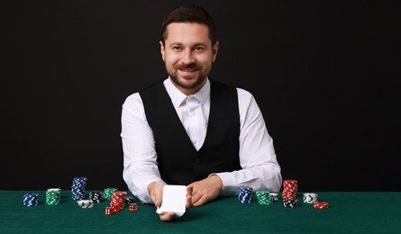 Professional croupier with playing cards at gambling table against black background