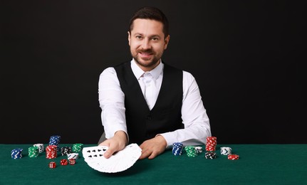 Professional croupier with playing cards at gambling table against black background