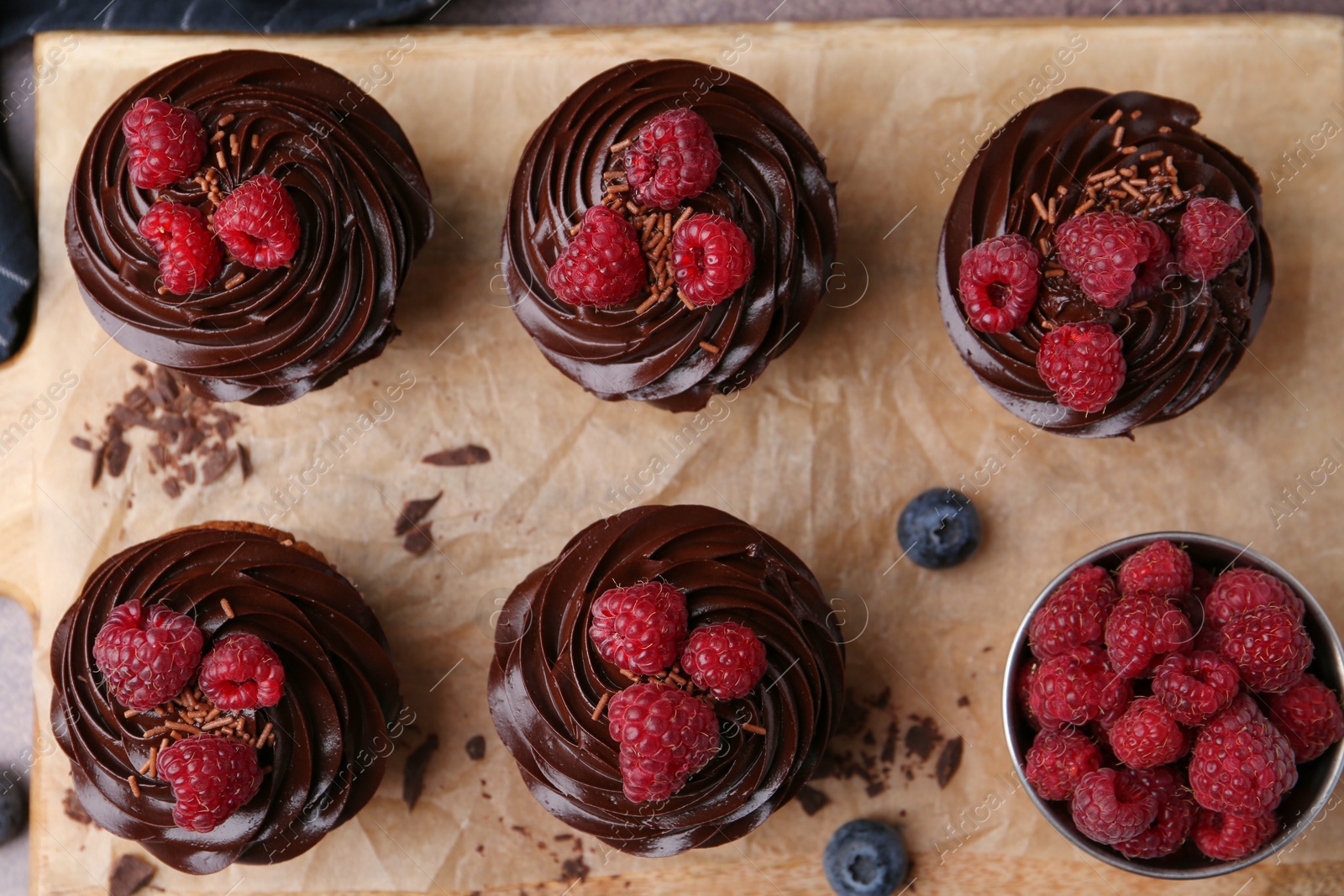 Photo of Tasty cupcakes with chocolate cream and raspberries on wooden board, flat lay