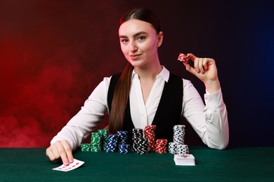 Photo of Professional croupier with casino chips and playing cards at gambling table on color background with smoke