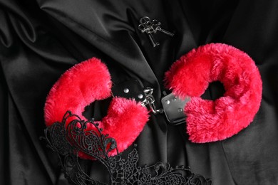 Photo of Red fluffy handcuffs, keys and lace mask on black fabric, flat lay