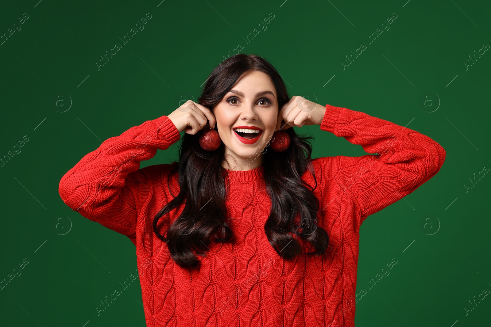 Photo of Happy woman trying Christmas balls as earrings on green background