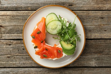 Photo of Delicious bagel with salmon, cream cheese, cucumber, avocado and pumpkin seeds on wooden table, top view