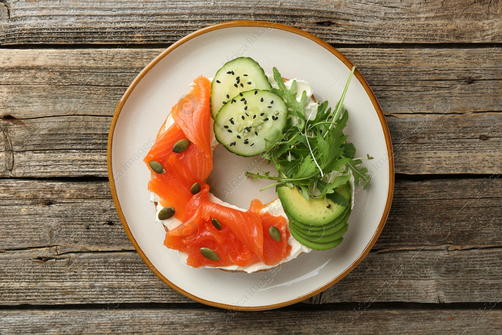 Photo of Delicious bagel with salmon, cream cheese, cucumber, avocado and pumpkin seeds on wooden table, top view