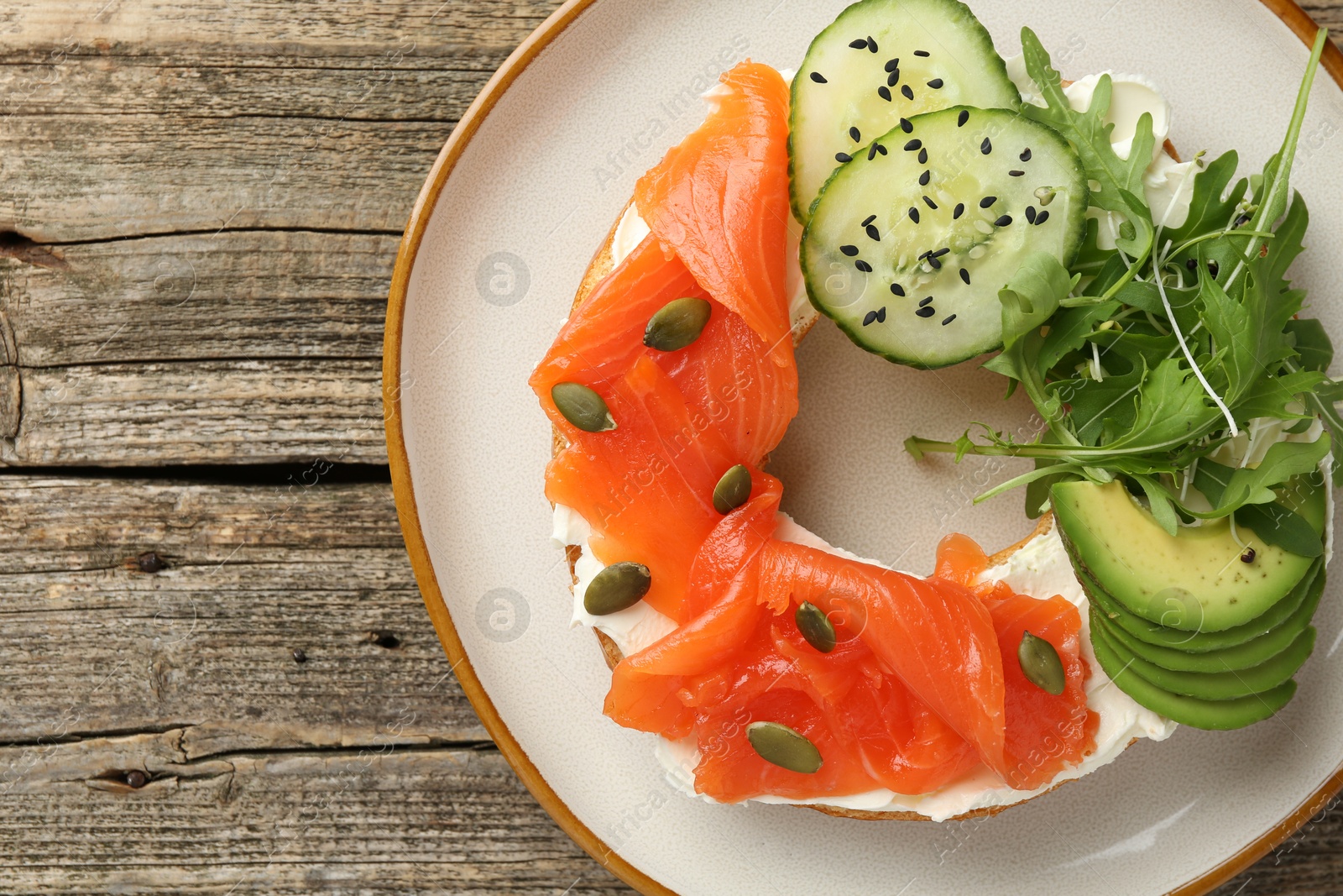 Photo of Delicious bagel with salmon, cream cheese, cucumber, avocado and pumpkin seeds on wooden table, top view. Space for text