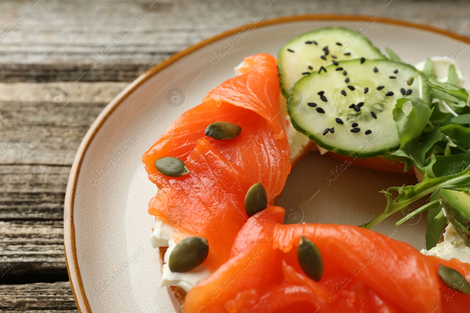 Photo of Delicious bagel with salmon, cream cheese, cucumber, avocado and pumpkin seeds on wooden table, closeup