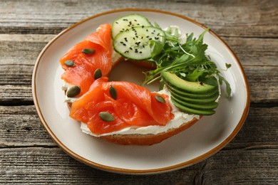 Photo of Delicious bagel with salmon, cream cheese, cucumber, avocado and pumpkin seeds on wooden table, closeup