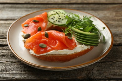 Photo of Delicious bagel with salmon, cream cheese, cucumber, avocado and pumpkin seeds on wooden table, closeup