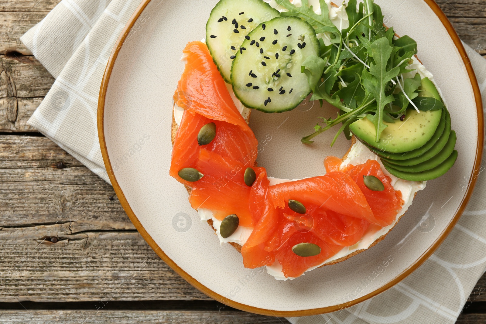 Photo of Delicious bagel with salmon, cream cheese, cucumber, avocado and pumpkin seeds on wooden table, top view