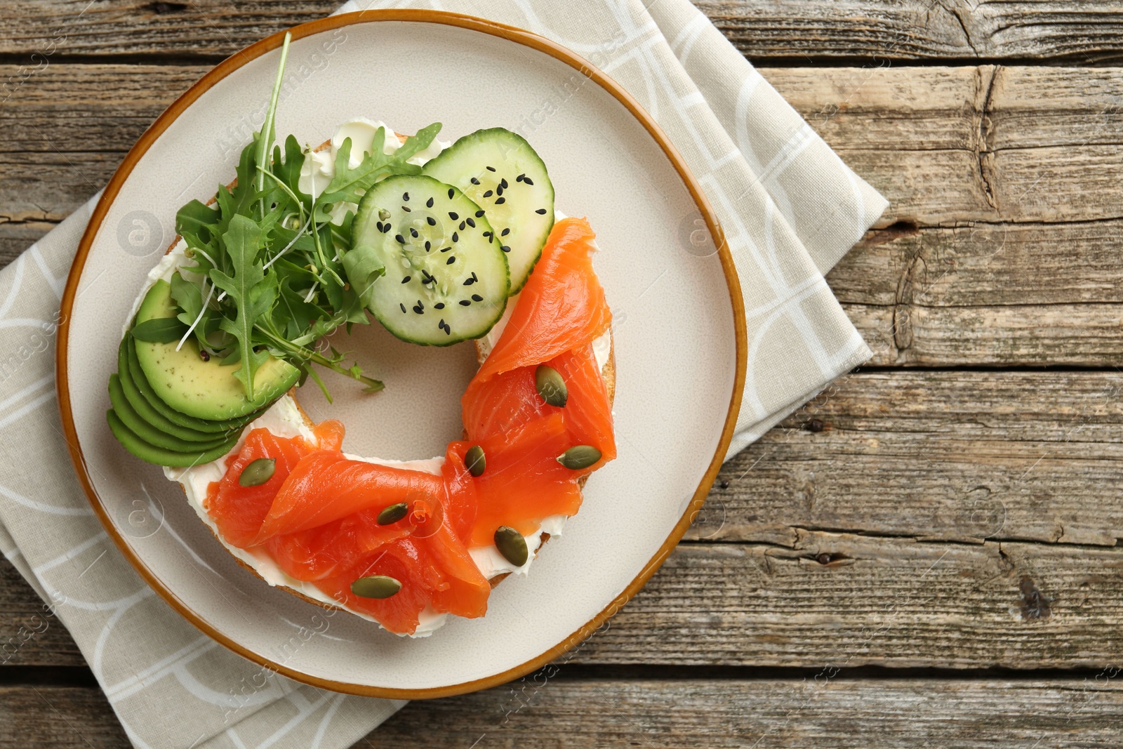 Photo of Delicious bagel with salmon, cream cheese, cucumber, avocado and pumpkin seeds on wooden table, top view. Space for text