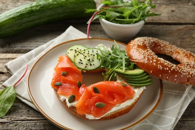 Photo of Delicious bagel with salmon, cream cheese, cucumber, avocado and pumpkin seeds on wooden table, closeup