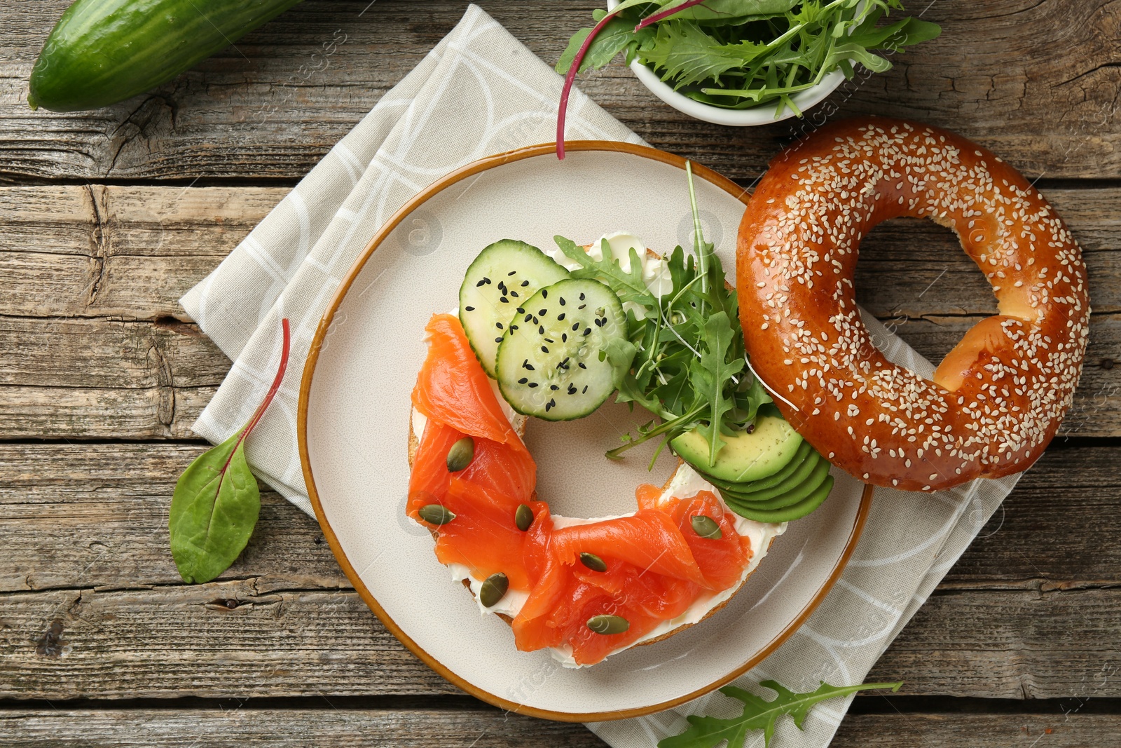 Photo of Delicious bagel with salmon, cream cheese, cucumber, avocado and pumpkin seeds on wooden table, flat lay