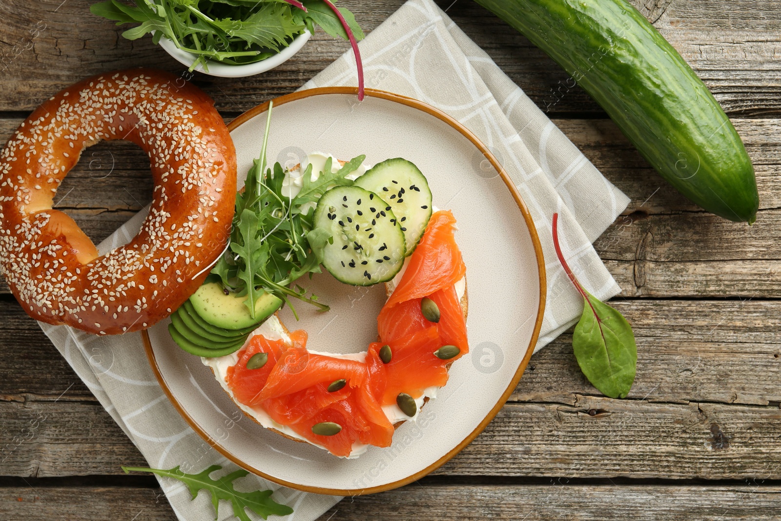 Photo of Delicious bagel with salmon, cream cheese, cucumber, avocado and pumpkin seeds on wooden table, flat lay