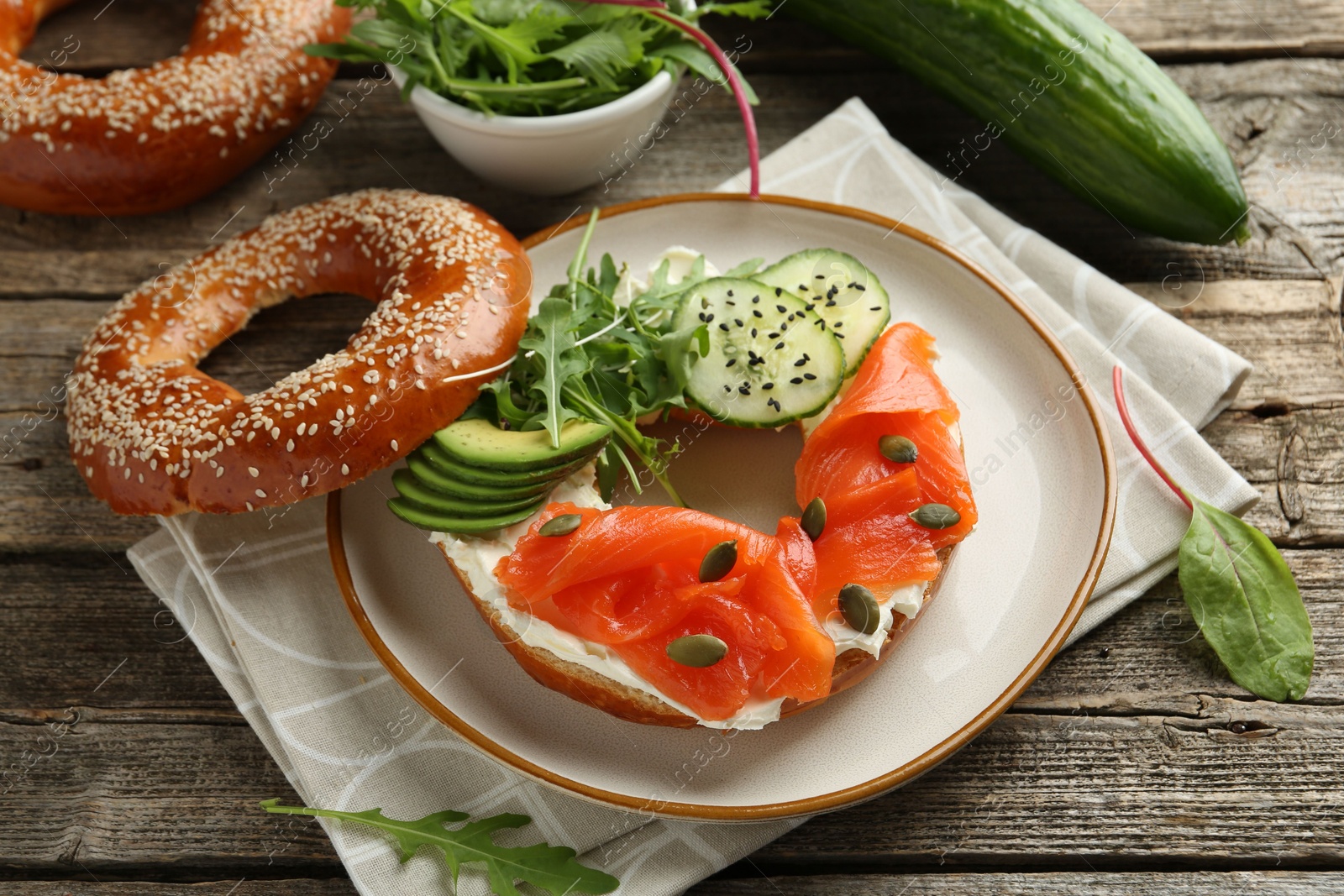 Photo of Delicious bagel with salmon, cream cheese, cucumber, avocado and pumpkin seeds on wooden table, closeup