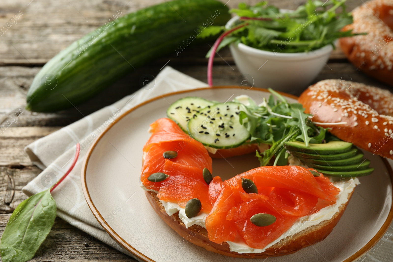 Photo of Delicious bagel with salmon, cream cheese, cucumber, avocado and pumpkin seeds on wooden table, closeup