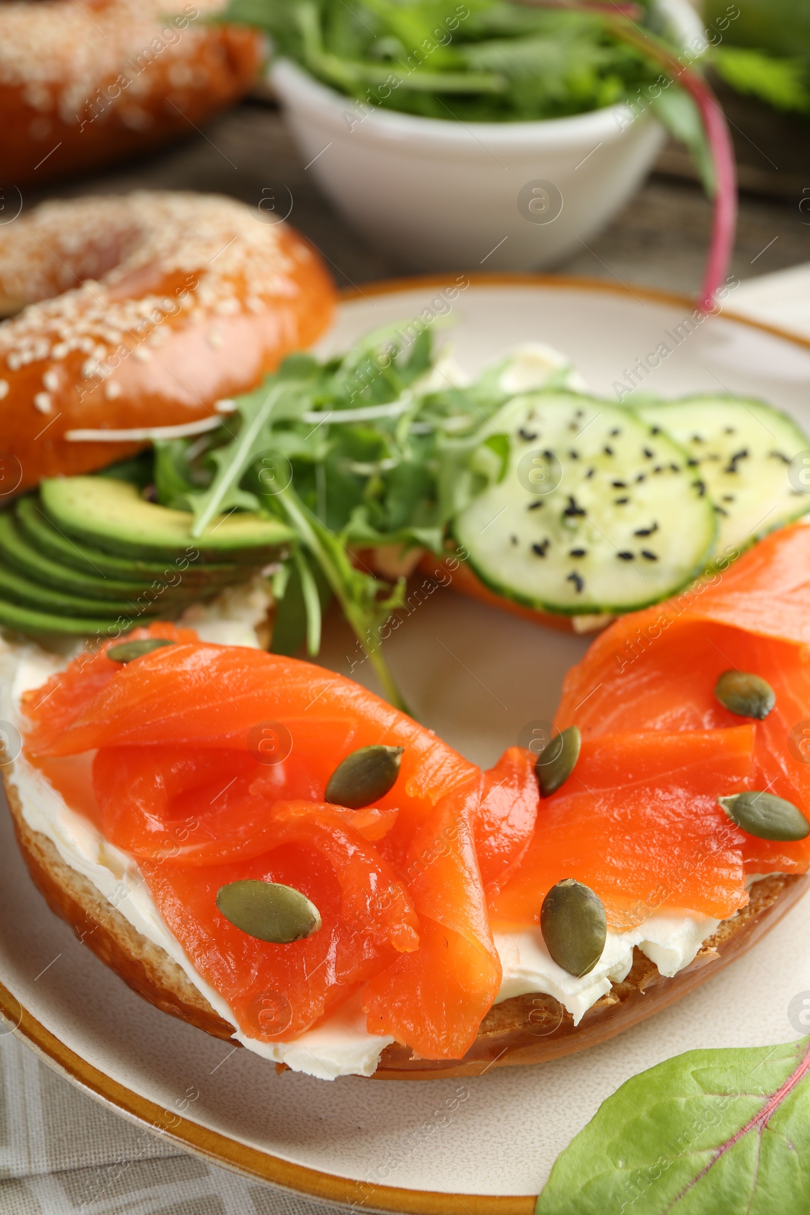 Photo of Delicious bagel with salmon, cream cheese, cucumber, avocado and pumpkin seeds on table, closeup