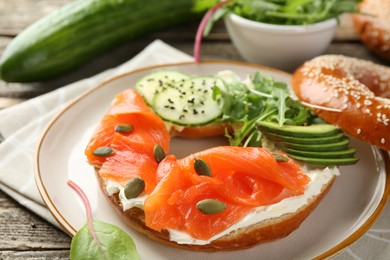 Photo of Delicious bagel with salmon, cream cheese, cucumber, avocado and pumpkin seeds on wooden table, closeup