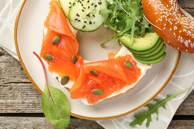 Photo of Delicious bagel with salmon, cream cheese, cucumber, avocado and pumpkin seeds on wooden table, top view