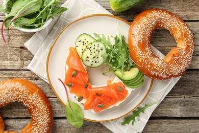 Photo of Delicious bagel with salmon, cream cheese, cucumber, avocado and pumpkin seeds on wooden table, flat lay