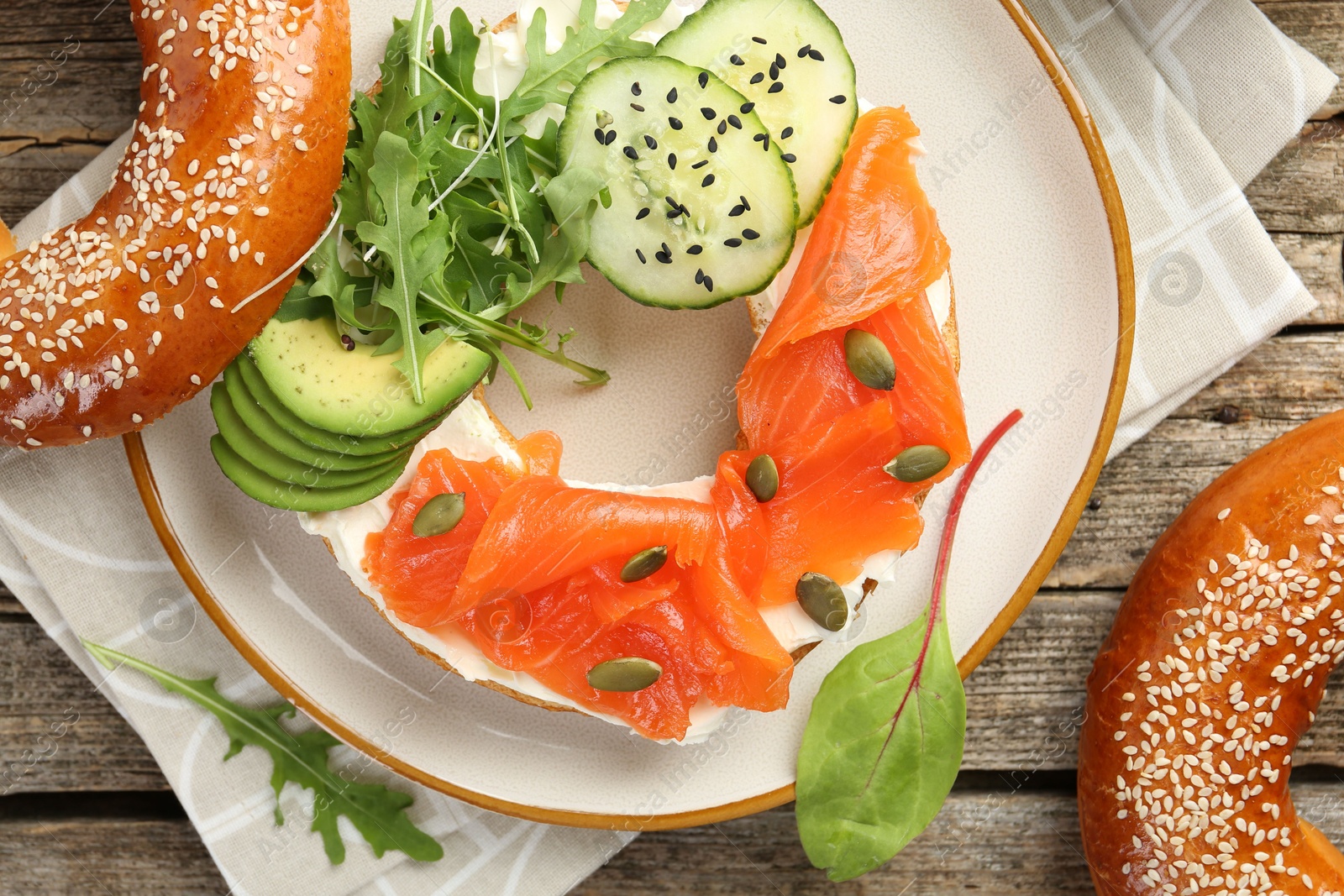 Photo of Delicious bagel with salmon, cream cheese, cucumber, avocado and pumpkin seeds on wooden table, flat lay