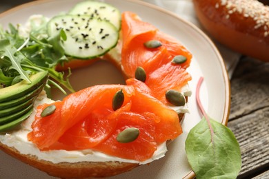 Photo of Delicious bagel with salmon, cream cheese, cucumber, avocado and pumpkin seeds on wooden table, closeup