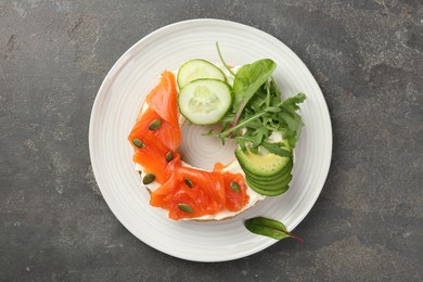 Photo of Delicious bagel with salmon, cream cheese, cucumber, avocado and pumpkin seeds on grey table, top view