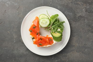 Photo of Delicious bagel with salmon, cream cheese, cucumber, avocado and pumpkin seeds on grey table, top view