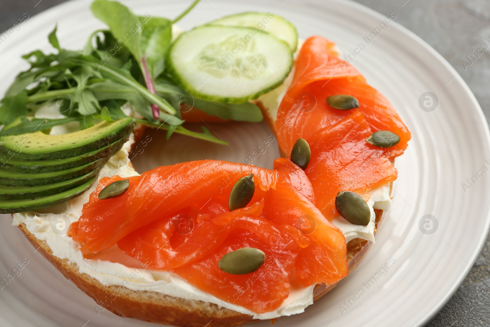 Photo of Delicious bagel with salmon, cream cheese, cucumber, avocado and pumpkin seeds on table, closeup