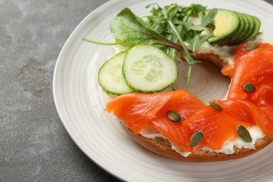 Photo of Delicious bagel with salmon, cream cheese, cucumber, avocado and pumpkin seeds on grey table, closeup