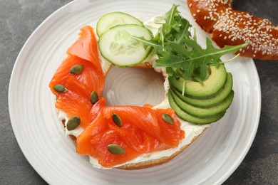 Photo of Delicious bagel with salmon, cream cheese, cucumber, avocado and pumpkin seeds on grey table, above view