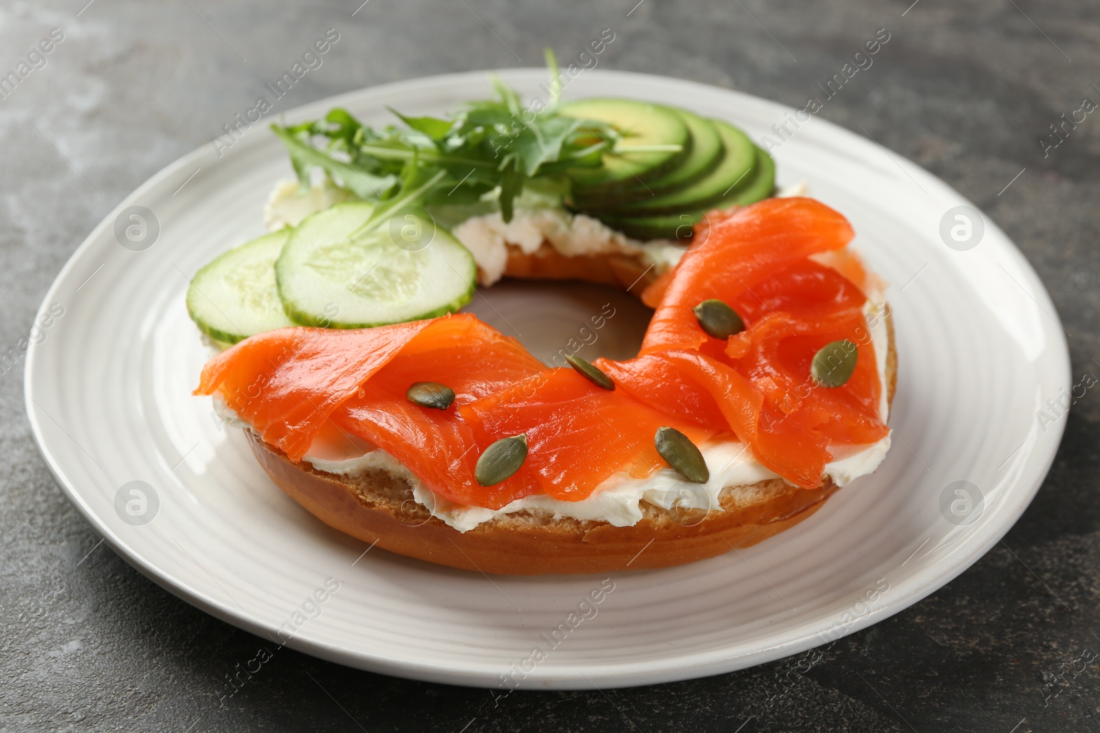 Photo of Delicious bagel with salmon, cream cheese, cucumber, avocado and pumpkin seeds on grey table, closeup