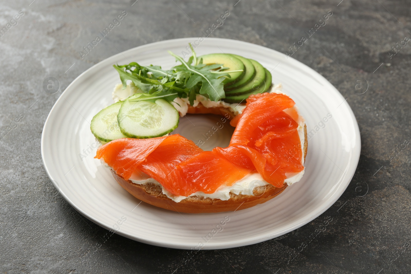 Photo of Delicious bagel with salmon, cream cheese, cucumber and avocado on grey table, closeup