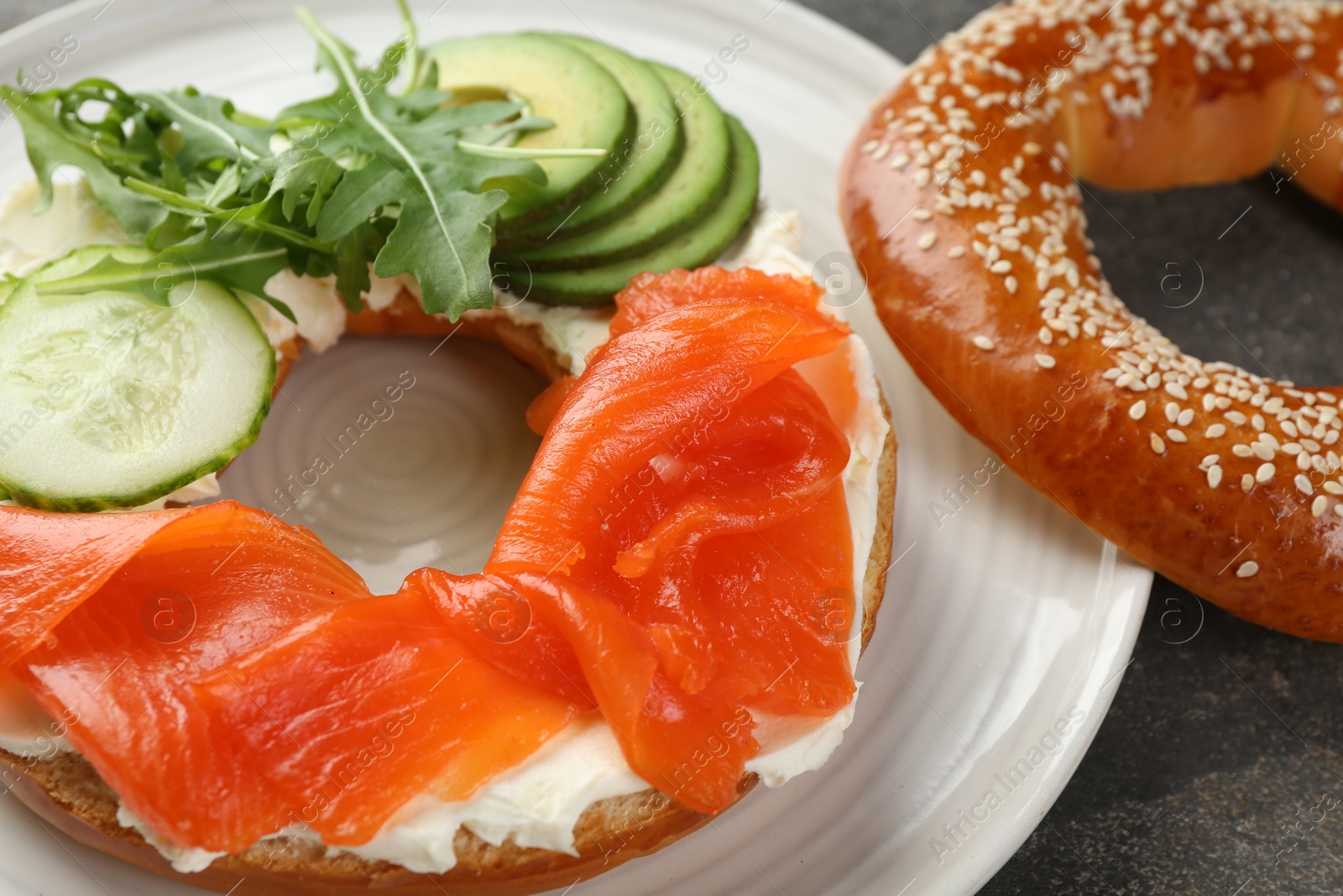 Photo of Delicious bagel with salmon, cream cheese, cucumber and avocado on grey table, closeup