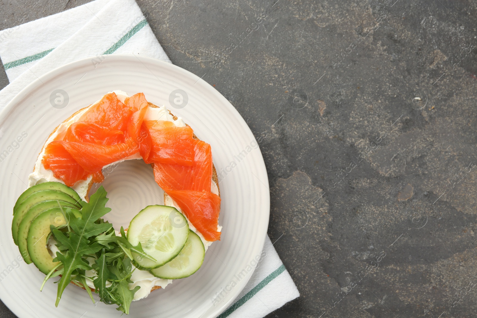 Photo of Delicious bagel with salmon, cream cheese, cucumber and avocado on grey table, top view. Space for text