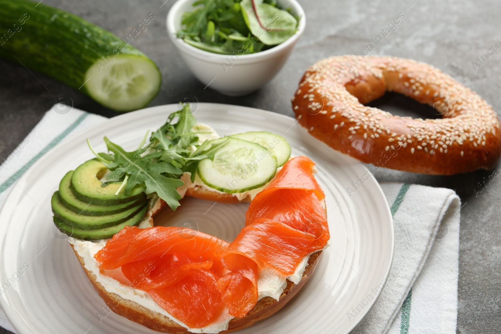 Photo of Delicious bagel with salmon, cream cheese, cucumber and avocado on grey table, closeup