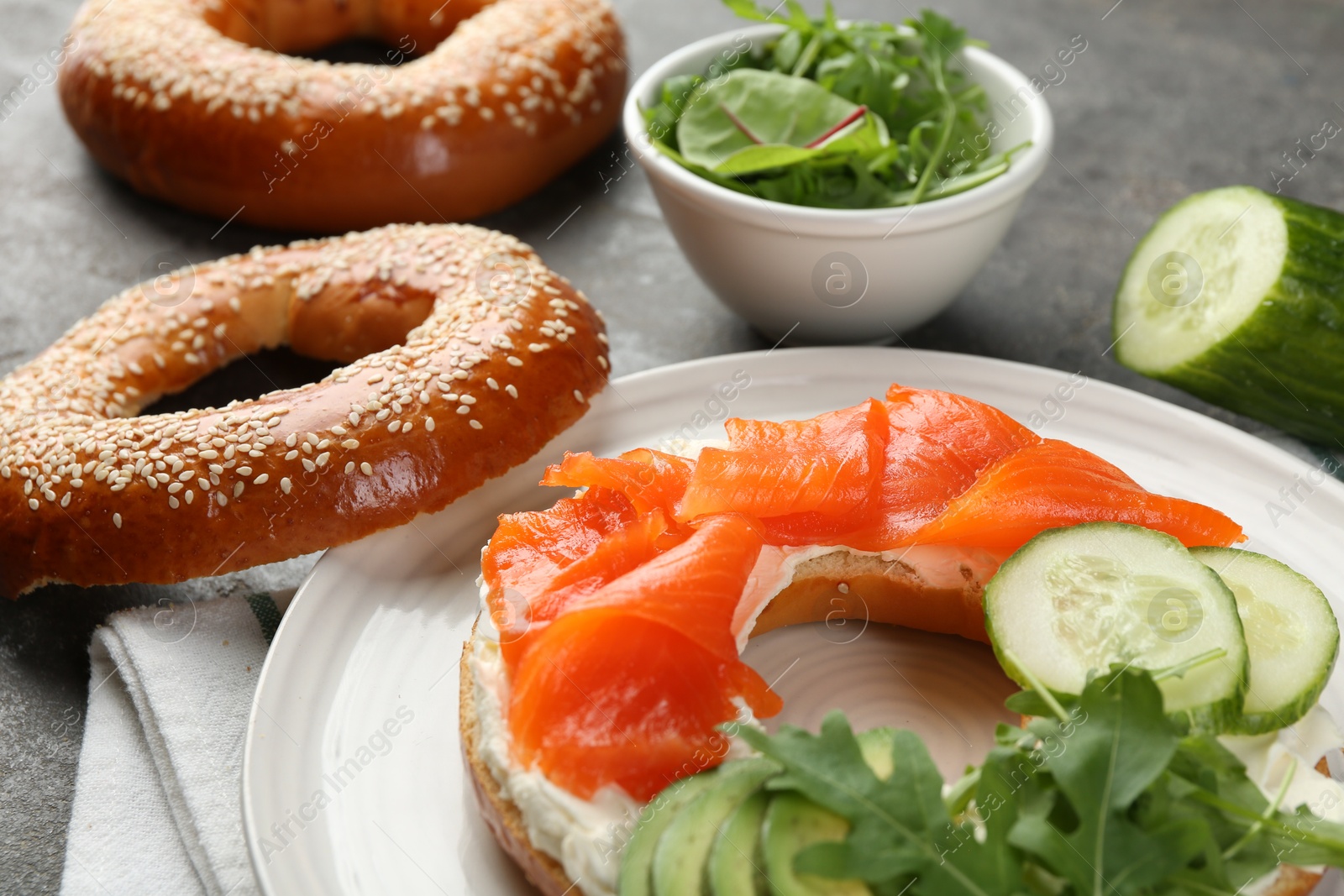 Photo of Delicious bagel with salmon, cream cheese, cucumber and avocado on grey table, closeup