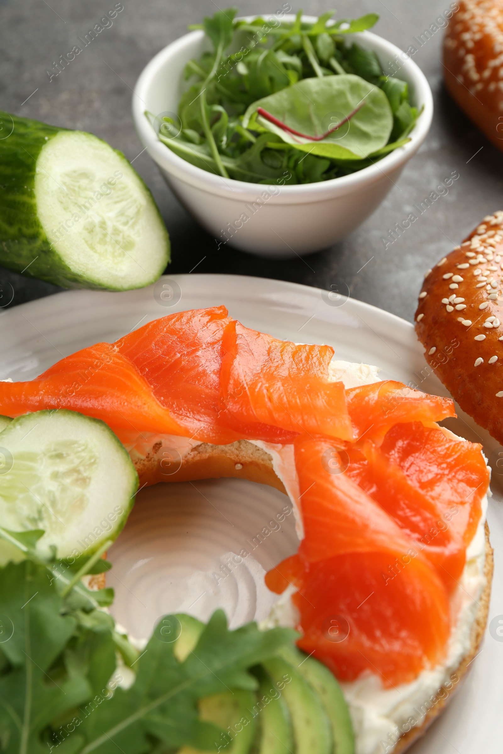 Photo of Delicious bagel with salmon, cream cheese, cucumber and avocado on grey table, closeup