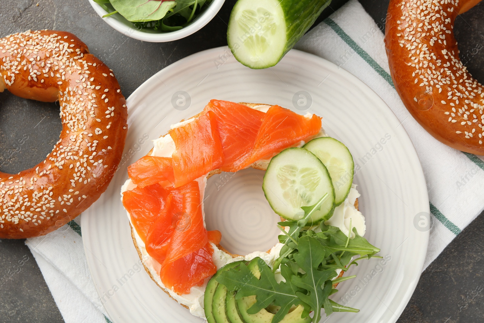 Photo of Delicious bagel with salmon, cream cheese, cucumber, arugula and avocado on grey table, flat lay