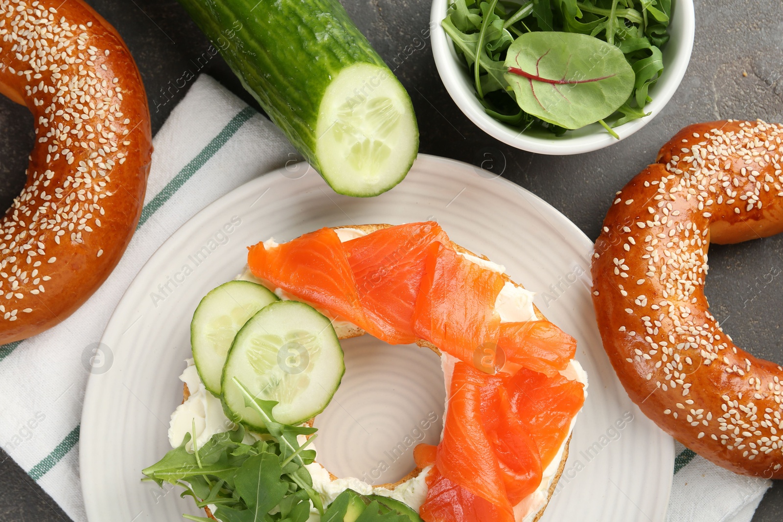 Photo of Delicious bagel with salmon, cream cheese, cucumber and arugula on grey table, flat lay