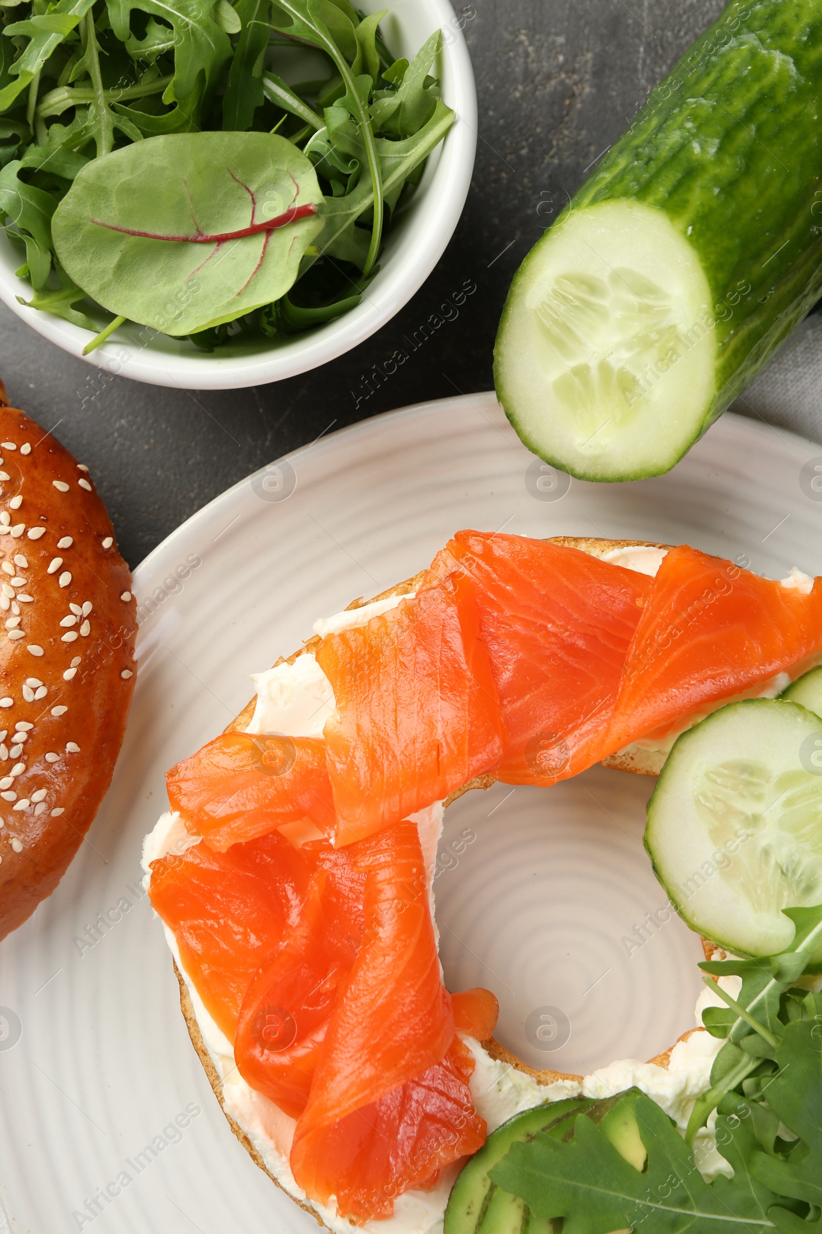 Photo of Delicious bagel with salmon, cream cheese, cucumber, arugula and avocado on grey table, flat lay