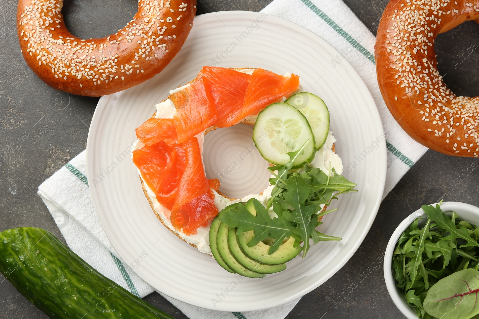 Photo of Delicious bagel with salmon, cream cheese, cucumber, arugula and avocado on grey table, flat lay