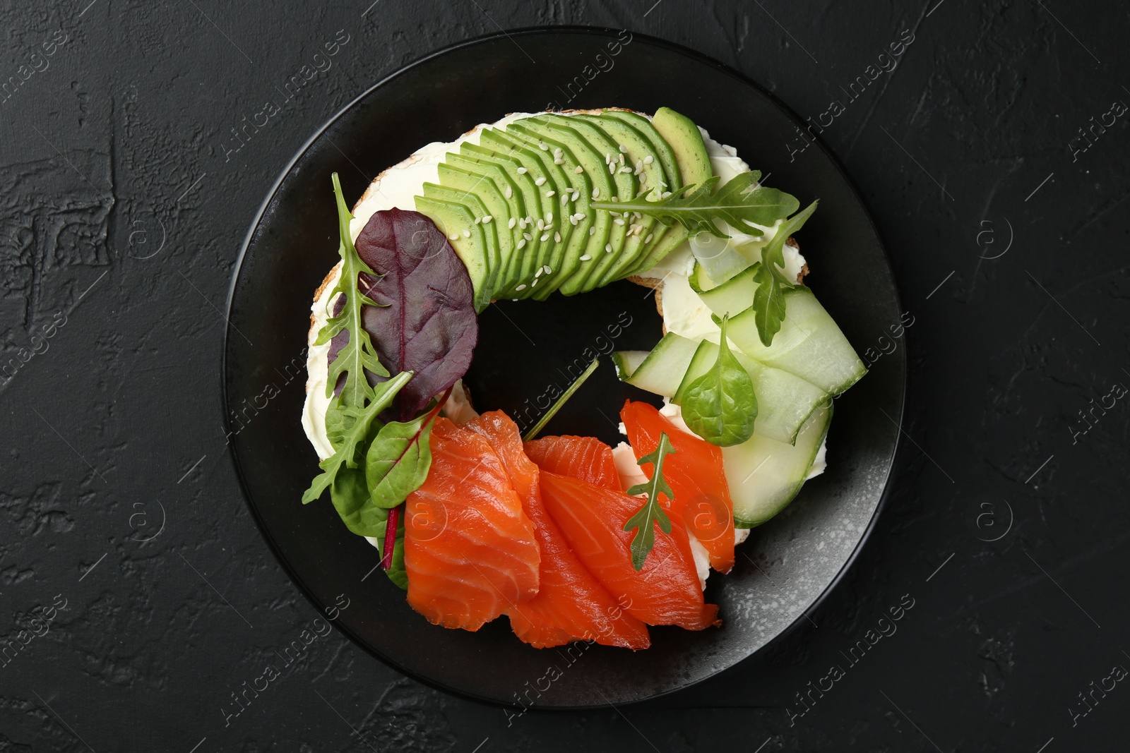 Photo of Delicious bagel with salmon, cream cheese, cucumber and avocado on black table, top view