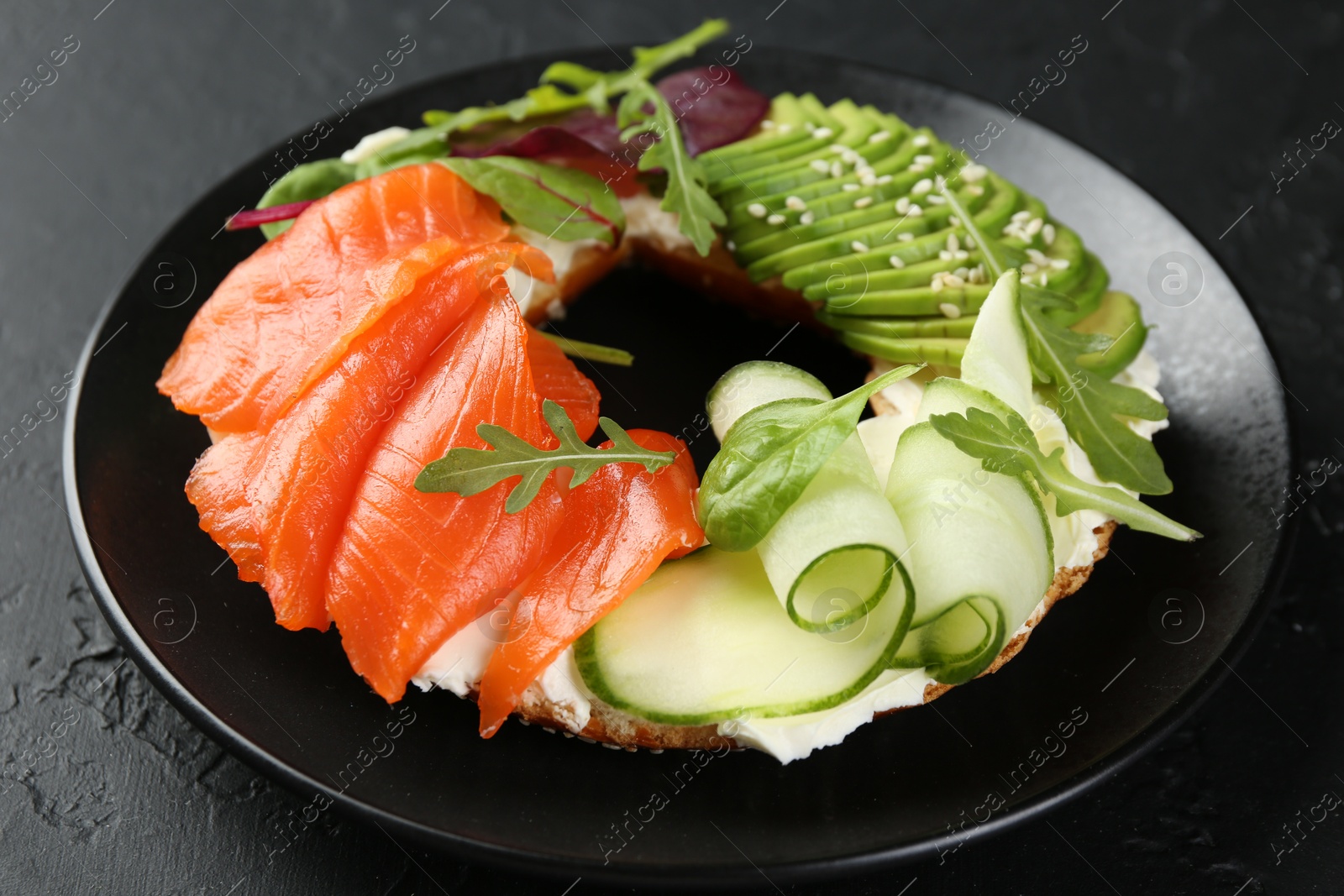 Photo of Delicious bagel with salmon, cream cheese, cucumber and avocado on black table, closeup