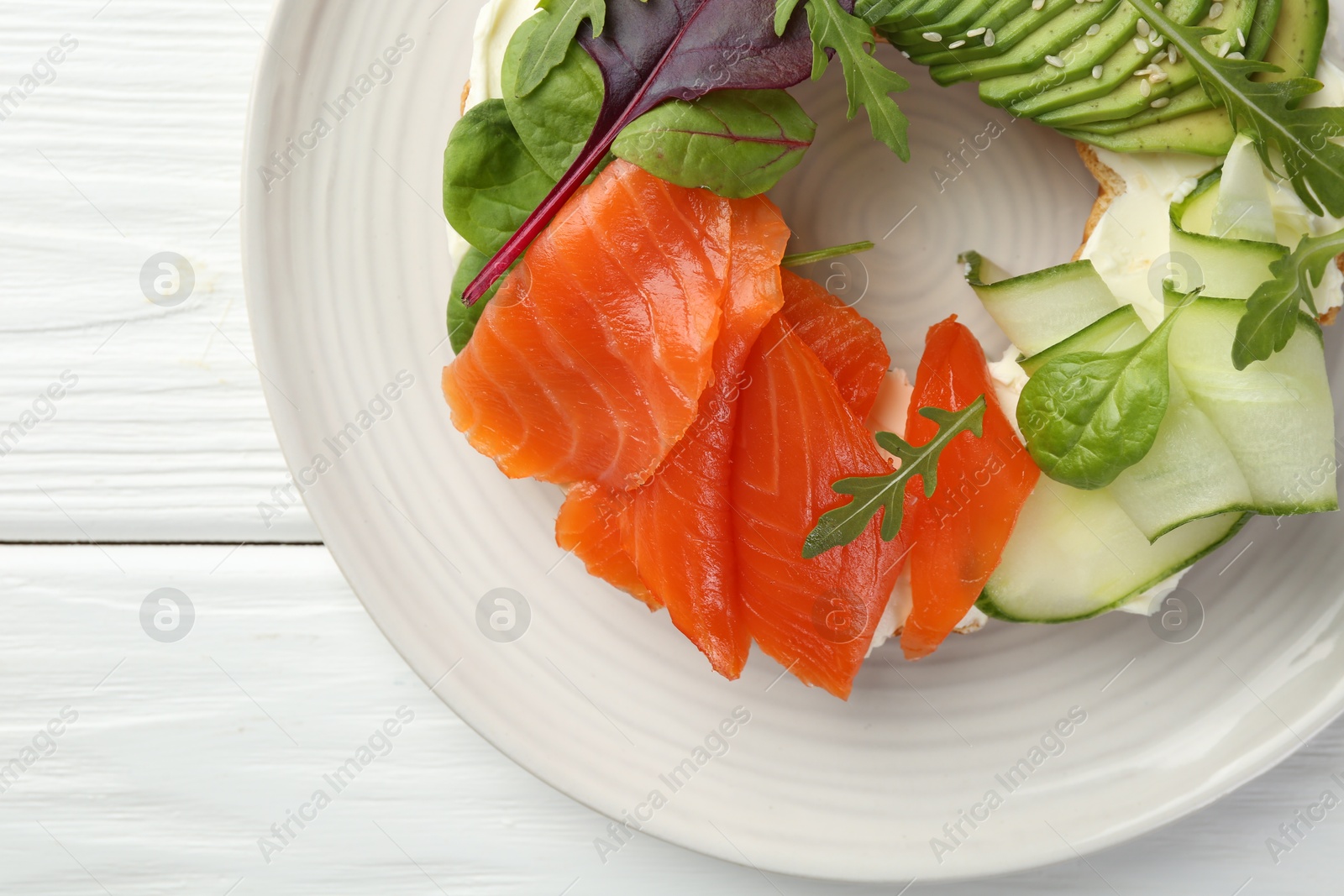 Photo of Delicious bagel with salmon, cream cheese, cucumber and avocado on white wooden table, top view