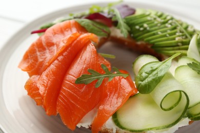 Photo of Delicious bagel with salmon, cream cheese, cucumber and avocado on white wooden table, closeup