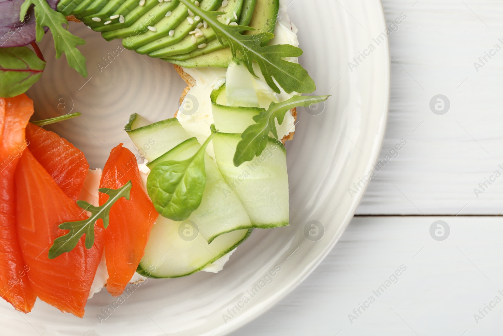 Photo of Delicious bagel with salmon, cream cheese, cucumber and avocado on white wooden table, top view