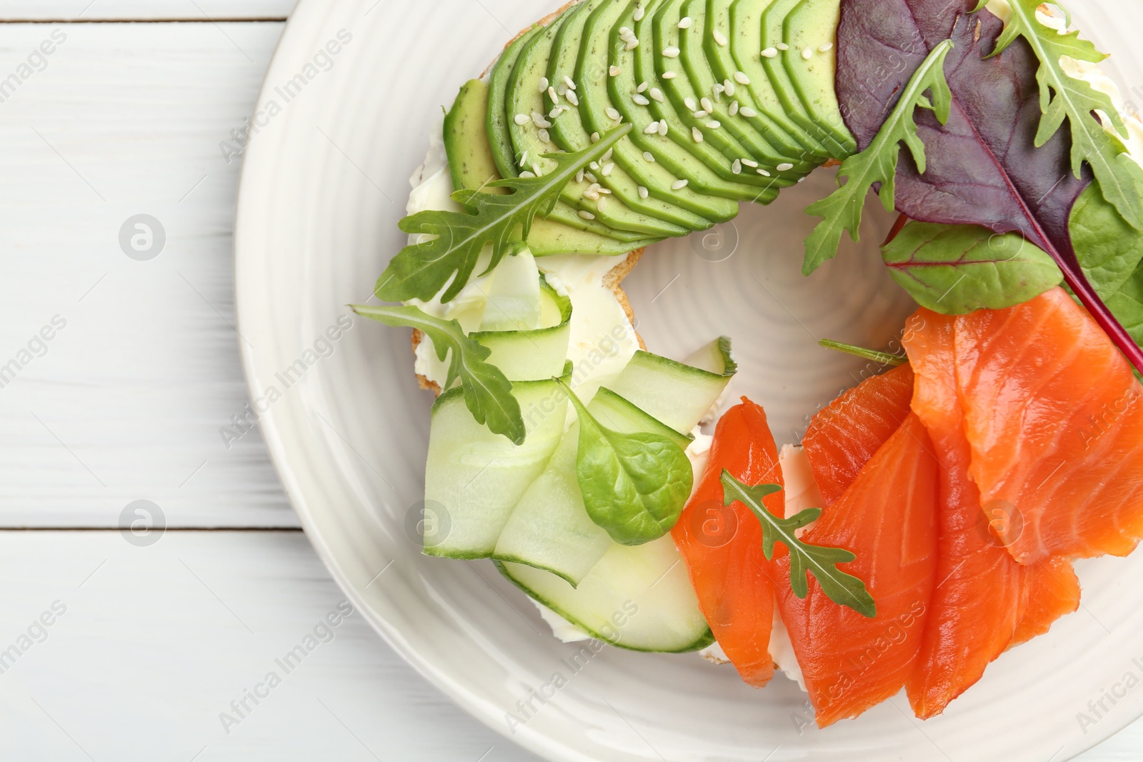 Photo of Delicious bagel with salmon, cream cheese, cucumber and avocado on white wooden table, top view