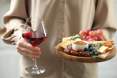 Photo of Woman holding board with different types of delicious cheese, other snacks and wine indoors, closeup