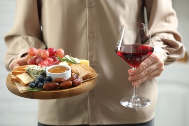 Photo of Woman holding board with different types of delicious cheese, other snacks and wine indoors, closeup