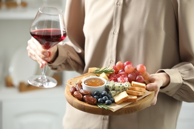 Photo of Woman holding board with different types of delicious cheese, other snacks and wine indoors, closeup